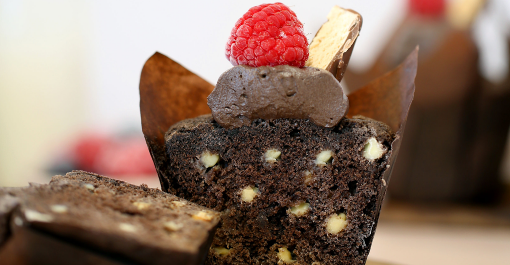 A chocolate muffin that shows an even distribution of chocolate chips, topped with buttercream and a raspberry