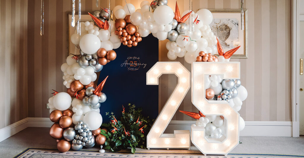 Decorative photo from the 25th Anniversary event, balloon arch with copper, white and silver balloons, light up 25 numbers and a blue board which has Kudos Blends 25th Anniversary celebration written on it.